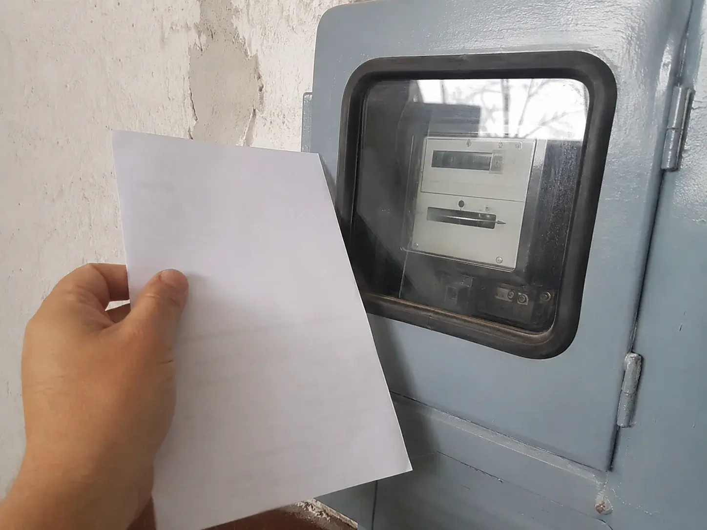 Električno brojilo na zidu, ruka drži papir za poređenje potrošnje