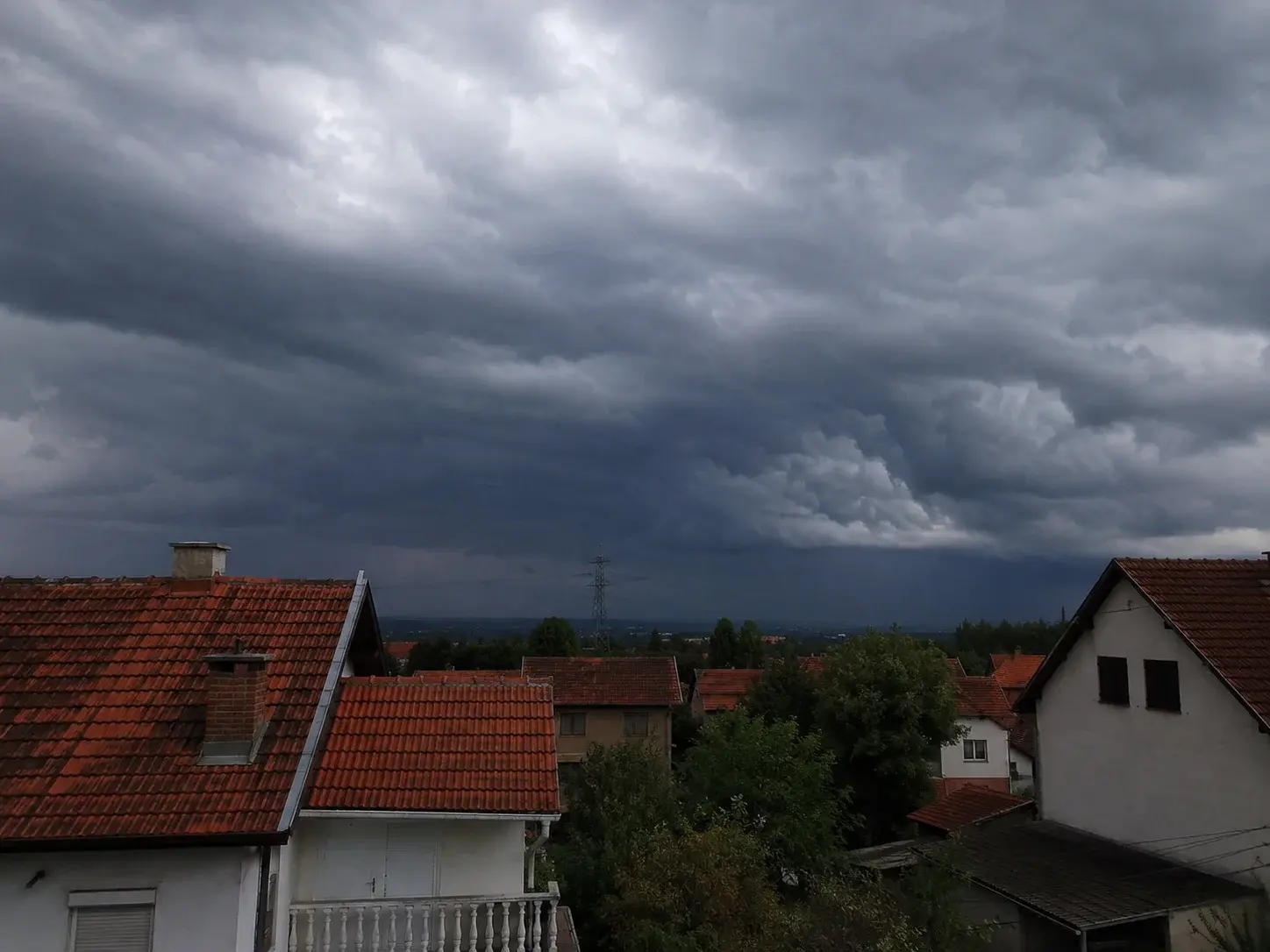 Olujno nebo nad krovovima kuća sa dalekovodnim stubom u daljini